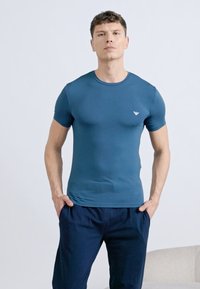 Νέος άνδρας στέκεται με τα χέρια στις τσέπες, φορώντας εφαρμοστό μπλε t-shirt και σκούρο μπλε παντελόνι, μπροστά σε απλό φωτεινό φόντο.