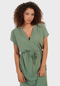 Robe verte en tissu doux, dotée d'un décolleté en V profond, de manches courtes, d'une taille élastique avec cordon de serrage, et de accents dorés.