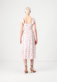 Henkilö, jolla on lyhyet vaaleat hiukset, pukeutuneena valkoiseen midi-mekkoon, jossa on vaaleanpunaisia kukkakuoseja, ja ruskeisiin korkokenkiin, seisoo selin.