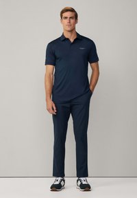 Tummansininen polo-paita lyhyillä hihoilla ja kontrastivärinen kaulus, yhdistettynä tummansinisiin housuihin ja urheilukengiin, joissa on valkoiset nauhat ja kumipohjat.