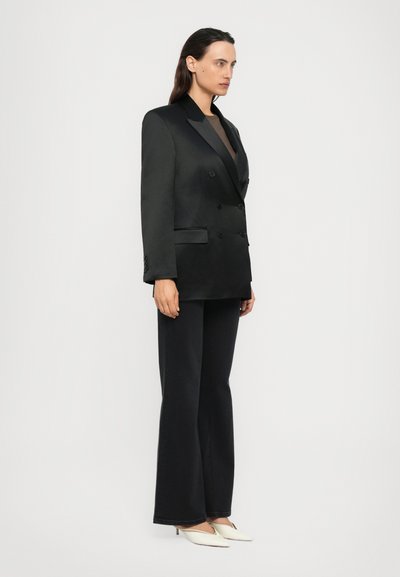 Vrouw staat met een dubbelzijdige zwarte blazer, zwarte wijde broek en witte puntige hakken tegen een eenvoudige lichte achtergrond.