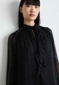 Schwarze transparente Bluse mit kleinen weißen Punkten, hohem Kragen, Schleifenverschluss, Puffärmeln und subtilen Rüschendetails an der Front.
