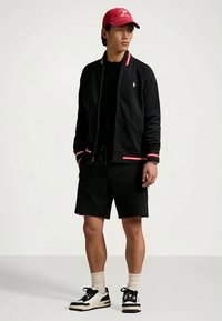 Melna džinsa jaka ar rāvējslēdzi, sarkanu un baltu svītrojumu aprocēs, melnas šorti, baltas un melnas krāsas kurpes un sarkana cepure. Vienkāršs, ikdienišķs apģērbs.