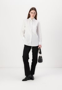 White striped button-up shirt with pocket, paired with black tailored pants and black heeled boots. Holds a small black bag with a strap.