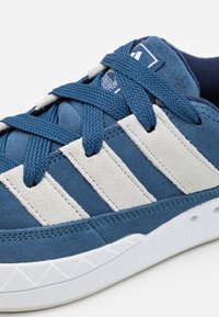 Sininen mokkajalkine valkoisilla mokkanauhoilla, teksturoidulla päällisellä ja sinisillä nauhoilla. Varusteena valkoinen kumipohja, jossa on tekstuuroitu kuvio.