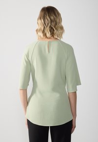 Top verde chiaro con maniche corte e orlo arrotondato, caratterizzato da un piccolo dettaglio a occhio dietro. Tessuto testurizzato con un motivo sottile.