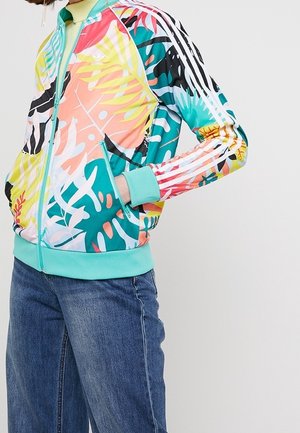 Personne portant une veste zippée à imprimé tropical coloré avec des poignets turquoise et un jean bleu, les mains dans les poches de la veste.