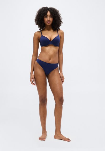 Femme aux cheveux bouclés, debout pieds nus, portant un soutien-gorge et une culotte assortis bleu marine, sur un fond clair uni.