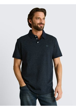 Homme barbu aux cheveux bruns porte un polo bleu foncé à manches courtes et un jean bleu, debout avec une main dans la poche, souriant et regardant vers la droite.