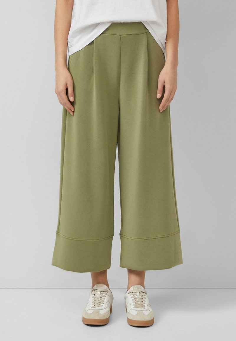 Osoba ubrana w szerokie, oliwkowozielone spodnie o kroju cropped, białe trampki oraz białą koszulkę wpuszczoną do spodni na jednolitym szarym tle.