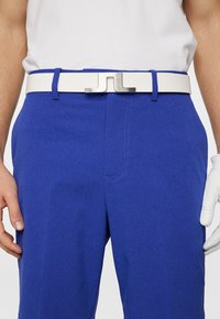 Blå teksturerede shorts med et hvidt bælte med en sølvspænde. En golfhandske er delvist synlig på højre hånd.