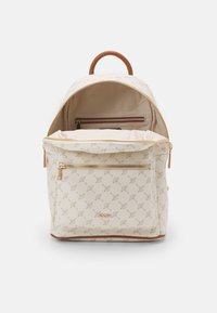 Béžový batoh se zlatými kovovými doplňky, s texturovaným vzorem, kapsami na zip a hnědým horním madlem. Viditelná vnitřní kapsa.