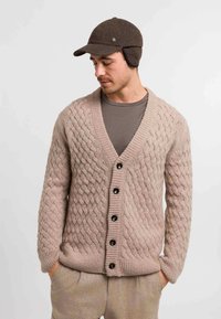 Beige cable-knit cardigan with a deep V-neck, six dark buttons, and ribbed cuffs. Worn over a gray T-shirt, paired with light trousers.