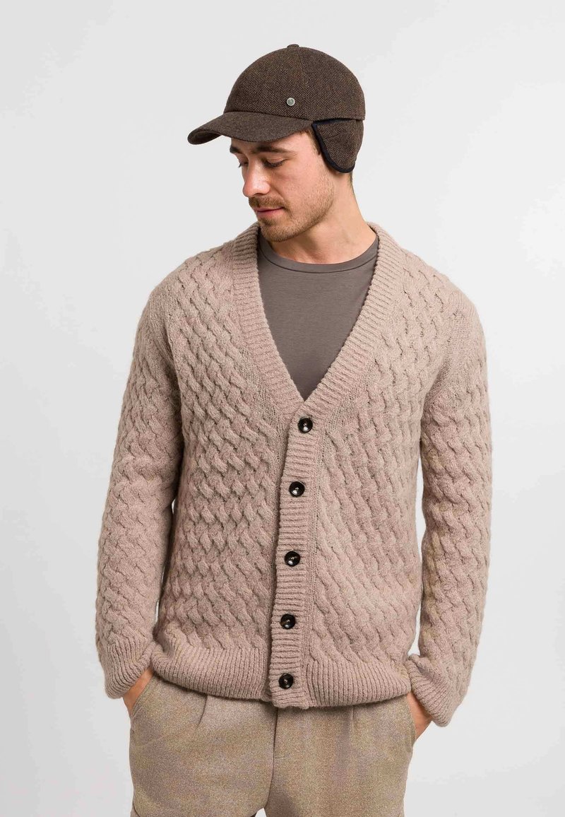 Beige cable-knit cardigan with a deep V-neck, six dark buttons, and ribbed cuffs. Worn over a gray T-shirt, paired with light trousers.