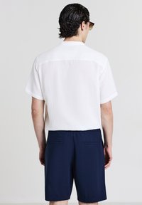 Uomo con capelli scuri che indossa occhiali da sole, una camicia bianca a maniche corte e pantaloni corti blu navy, in piedi contro uno sfondo chiaro e uniforme.