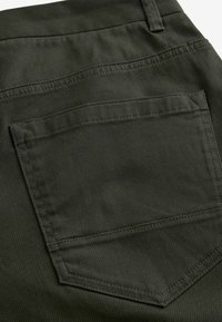 Poche arrière en tissu vert olive avec une finition texturée, présentant des coutures doubles et un bord supérieur légèrement incurvé.