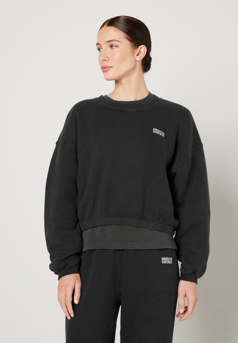 Black oversized sweatshirt with a ribbed hem and cuffs. Features a small white "American Vintage" logo on the chest. Classic crew neck design.