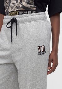 Personne portant un pantalon de jogging gris clair avec un cordon noir et un logo VD avec des ailes sur la cuisse gauche, associé à un t-shirt graphique noir.