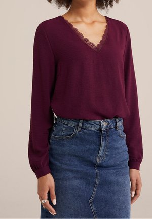 Femme portant un haut prune à manches longues avec un col en V orné de dentelle, associé à une jupe en jean bleue taille haute.