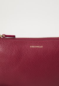 Red leather clutch with a textured surface, featuring a gold zipper and embossed brand logo. Compact rectangular shape.