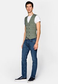 Zielona kamizelka w kratę z podwójnymi guzikami na białej koszulce, połączona z niebieskimi dżinsami i czarnymi sneakersami. Luźny fason, teksturowany materiał.