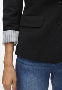 Svart blazer med en enkel ficka, med en rullad manschett av ljusblått tyg med vertikala ränder. Bärs med blå denimjeans.