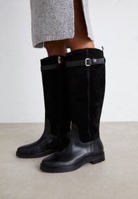 Schwarze kniehohe Stiefel aus einer Kombination von glattem Leder und Wildleder, mit einer silbernen Schnallenakzentuierung und kontrastierenden Texturen.