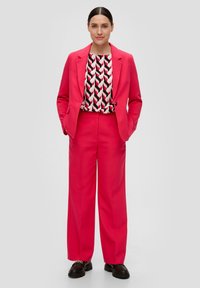 Un costume rose vif avec une veste cintrée et un pantalon à jambes larges, associé à un haut à motifs noir, rouge et blanc. Des chaussures noires épaisses.