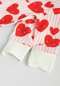 Tessuto a motivo di cuori rossi con volti sorridenti su uno sfondo a righe rosa chiaro e bianco; polsini bianchi a coste alla fine delle maniche.