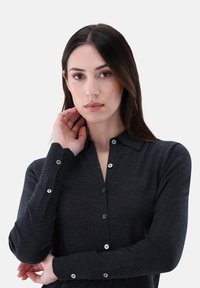 Dunkelgraues Langarmshirt mit Button-Down-Kragen, ausgestattet mit silbernen Knöpfen und einem weichen, strukturierten Stoff. Modell berührt leicht ihren Hals.
