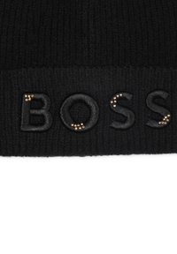 Beanie noir en tricot avec un logo "BOSS" brodé et des accents dorés. Texture côtelée, matériau doux et design simple.