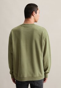 Man die met zijn rug naar de camera draait en een olijfgroene sweatshirt draagt met geribbelde manchetten en zoom, gecombineerd met donkergrijze jeans tegen een eenvoudige achtergrond.