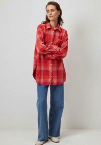Raudona languota flanelinė marškiniai su mygtukų užsegimu, ilgomis rankovėmis ir šoninėmis įpjovomis, derinami su šviesiai mėlynomis džinsais ir kreminėmis spalvos bateliais.