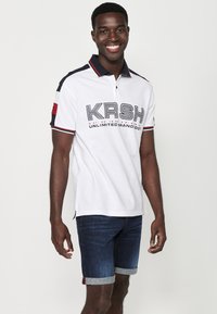 Bijela polo majica s crnim i crvenim detaljima, na kojoj se ističe tekst "KRSH" i "UNLIMITED BRAND" u debelom fontu. Uparena s traper šorcem.