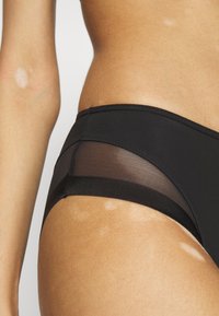 Black bikini bottom featuring sheer mesh panels, a smooth texture, and a low-rise design. Includes fine stitching along the edges.