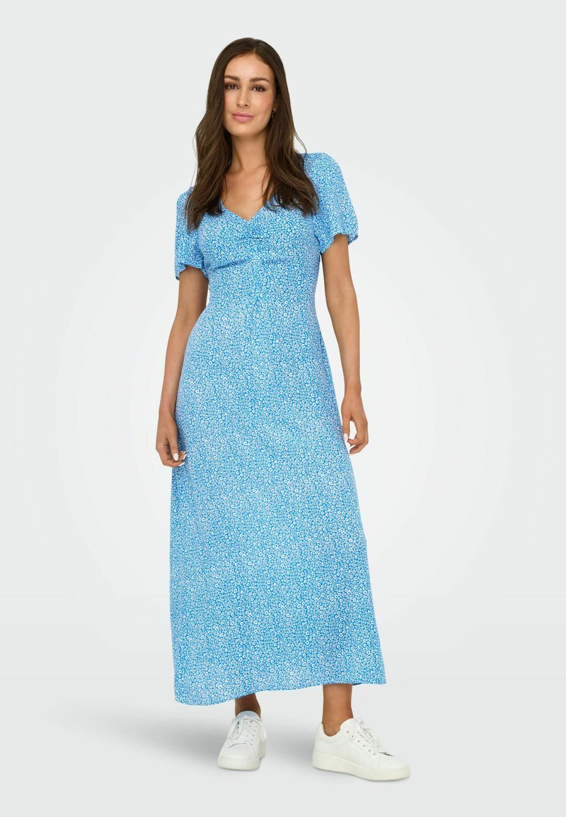 Femme aux longs cheveux bruns portant une robe longue bleue à fleurs à manches courtes et des baskets blanches, debout devant un fond clair uni.