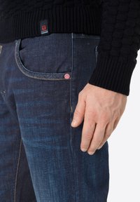 Donkerblauwe spijkerbroeken met zichtbare stiksels, voorzien van een roze accentknop en een gestructureerde zwarte trui met geribde manchetten.