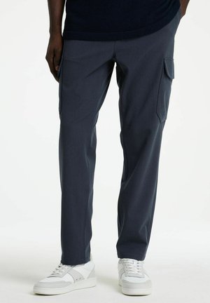 Pantalones cargo azul oscuro con bolsillos laterales con solapa, usados con zapatillas blancas de cordones, en una persona de pie.