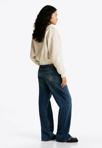 Creme cropped sweatshirt med ribbede manchetter, parret med brede jeans i blå med stjerner og studs på baglommen. Sorte sko.