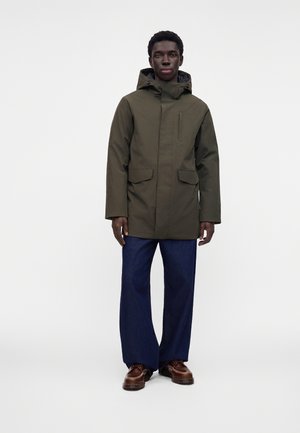 Homme portant un manteau à capuche vert foncé, un pantalon bleu à jambes larges et des chaussures en cuir marron, debout devant un fond clair uni.