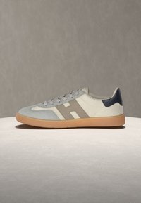 Παπούτσι τύπου sneaker χαμηλού προφίλ με μπεζ, γκρι και ναυτικόμπλε πάνελ, καφέ σόλα από καουτσούκ και ένα μεγάλο γκρι "H" στο πλάι.