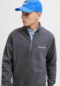 Sudadera gris con cremallera y textura suave, con bordado de la marca. Usada sobre una camisa a rayas azul clara y combinada con una gorra azul.