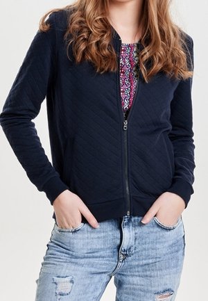 Femme portant une veste matelassée zippée bleu marine sur un haut à motifs colorés et un jean bleu clair déchiré, les mains dans les poches.