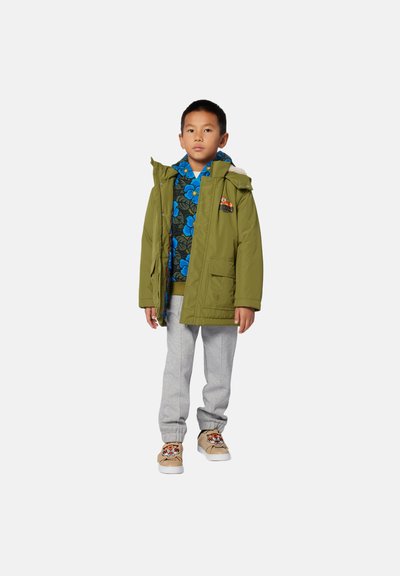 Enfant portant un manteau d'hiver à capuche vert olive par-dessus un pull floral bleu, un pantalon de survêtement gris et des baskets beiges avec un motif de visage de tigre, restant immobile.