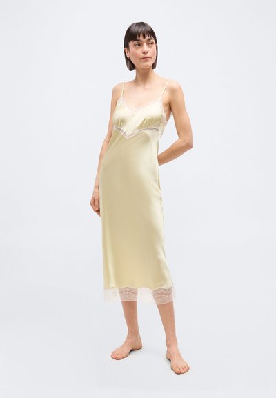 Γυναίκα που στέκεται ξυπόλητη, φορώντας ένα απαλό κίτρινο σατινένιο φόρεμα με δαντέλα στο μπούστο και το τελείωμα, κοιτάζοντας ελαφρώς προς τα αριστερά της.