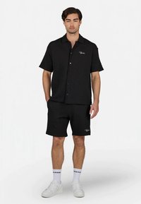 Man standing wearing black short-sleeve button shirt and matching shorts with white socks and white sneakers on white background.