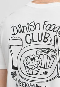 Balta marškinėliai su juodu grafikiniu piešiniu, vaizduojančiu maisto produktus, įskaitant keksiukus ir stiklą, pažymėti „Daniškos virtuvės KLUBAS“, su tekstu skirtingais dydžiais.
