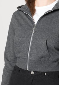 Sudadera gris corta con cremallera, con un bolsillo frontal y capucha. Lleva puesto una camisa blanca, acompañada de pantalones negros. Textura suave.