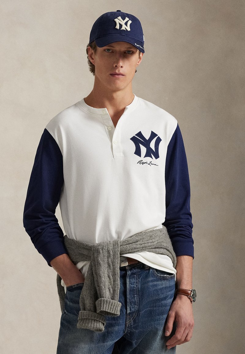Chemise henley blanche à manches bleu marine, avec un grand logo NY bleu marine. Pull en tricot torsadé gris noué autour de la taille. Jeans en denim décontractés.