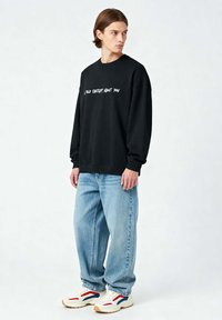 Felpa nera con testo bianco che dice "HO PARLATO CON CHATGPT DI TE." Abbinata a jeans baggy blu chiaro e sneakers bianche con dettagli rossi e blu.